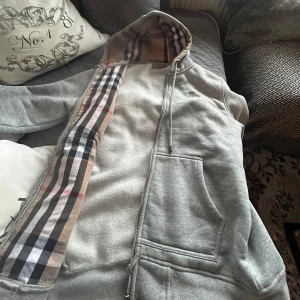 Grå  Burberry hoodie - Säljer en snygg grå hoodie från Burberry med klassiskt rutigt mönster i huvan. Den har en dragkedja framtill och en broderad detalj på bröstet. Perfekt för en stilren look. Skriv vid frågor 