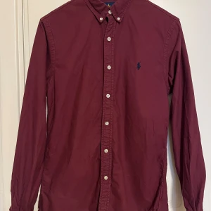 Vinröd skjorta från Ralph Lauren - Snygg vinröd skjorta från Ralph Lauren i slim fit. Skjortan har långa ärmar och knappar framtill samt en broderad logotyp på bröstet. Perfekt för en stilren look. Sparsamt använd i mycket gott skick. Se gärna mina andra annonser, så kan allt skickas i samma sändning. 