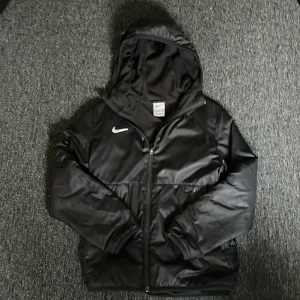 Svart jacka från Nike - Säljer en svart jacka från Nike med huva och dragkedja. Jackan har en stilren design med Nike-logga på bröstet. Perfekt för kyligare dagar.