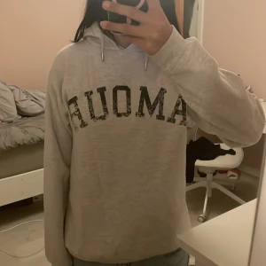 Grå hoodie från Gina Tricot - Säljer en grå hoodie från Gina Tricot med texten 'AMOUR' på framsidan och en stjärna på baksidan. Tröjan har en bekväm passform med långa ärmar och en klassisk huva. Perfekt för en simple outfit.