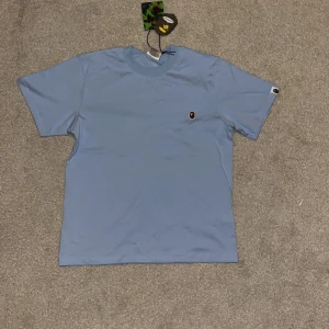 Blå t-shirt från A Bathing Ape - Säljer en snygg blå t-shirt från A Bathing Ape med det ikoniska apansikte-logotypen broderad på bröstet. T-shirten har en normal passform och korta ärmar. Perfekt för en stilren och avslappnad look.