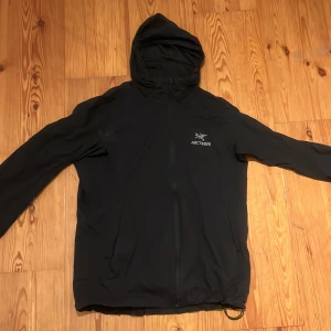 Svart vindjacka från Arc'teryx - Säljer en svart vindjacka från Arc'teryx med huva och dragkedja framtill. Jackan har en stilren design med Arc'teryx-loggan på bröstet. Perfekt för blåsiga dagar och utomhusaktiviteter. Använd en gång och har inga deffekter alls. Skriv gärna vid några funderingar