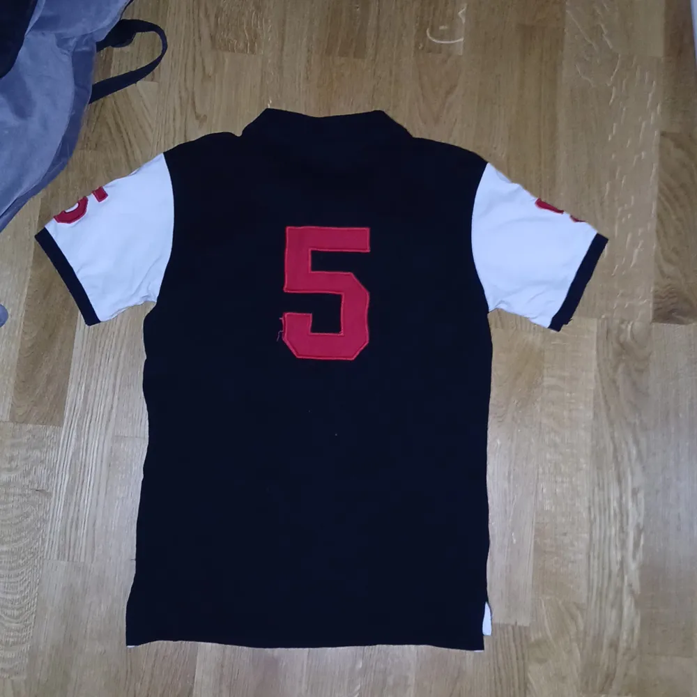 Snygg svart pikétröja från Ralph Lauren med vita ärmar och röda detaljer. Tröjan har en broderad logga och texten 'TOKYO' på framsidan samt en stor siffra '5' på baksidan. Perfekt för en stilren look. EXTRAPRIS I 1 DAG. T-paidat.