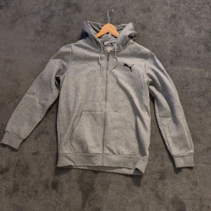 Grå huvtröja från Puma - Säljer en stilren grå huvtröja från Puma med dragkedja och kängurufickor. Tröjan har en justerbar huva med dragsko och ribbade muddar vid ärmslut och nederkant. Perfekt för en avslappnad look!