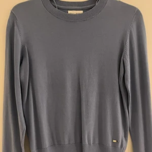 Blå tröja från Holly & Whyte/Lindex - Snygg blå tröja från Holly & Whyte by Lindex med ribbade detaljer och dekorativa guldknappar på ärmarna. Perfekt för en stilren look.