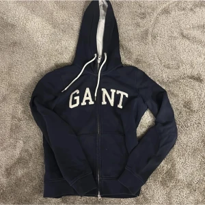 Mörkblå hoodie från GANT - Säljer en snygg mörkblå hoodie från GANT med dragkedja och vit text på bröstet. Den har en justerbar huva med vita snören och är perfekt för en avslappnad stil. Passar bra till både jeans och joggers.