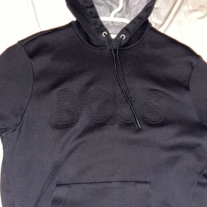 Svart hoodie från Boss - Säljer en stilren svart hoodie från Boss med broderad logga på bröstet. Den har en justerbar huva med snören och en praktisk magficka. Perfekt för en avslappnad look.