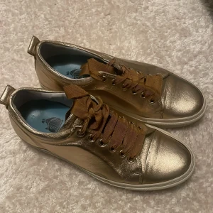 Guldiga sneakers från Lanvin - Snygga guldiga sneakers från Lanvin med breda bruna snören. Skorna har en glansig finish och en bekväm sula. Perfekta för att ge en touch av lyx till din outfit.