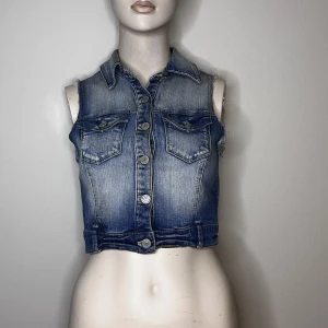 Blå croppad jeansväst från Gina tricot - Blå croppad jeansväst från Gina tricot. Västen har en klassisk krage och knappar framtill. Passar bra till både t-shirt och klänning.