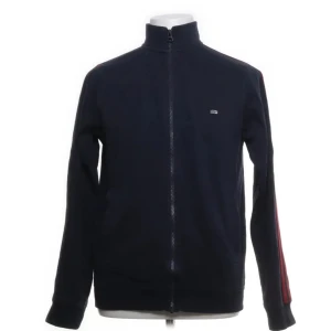 Mörkblå track jacket från Lexington - Säljer en stilren mörkblå track jacket från Lexington med dragkedja och röda ränder på ärmarna. Jackan är tillverkad i en mjuk bomullsblandning med nylon för extra komfort. Perfekt för en avslappnad look.