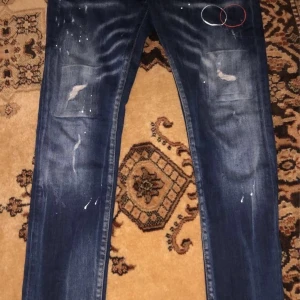Mörkblå jeans med slitningar från Dsquared2 - Snygga mörkblå jeans från Dsquared2 med coola slitningar och färgstänk för en trendig look. De har en klassisk femficksdesign och en bekväm passform. Perfekta för att ge din outfit en edgy touch.