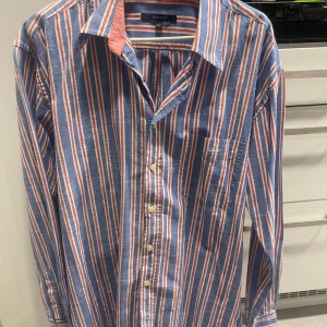 Randig skjorta från GANT - Säljer en snygg randig skjorta från GANT i blått, vitt och rött. Skjortan har långa ärmar och klassisk krage med knappar framtill. Perfekt för en stilren look. Osäker om de två knapparna ska vara där, och sista bilden ränderna är inte gula det är ljuset.