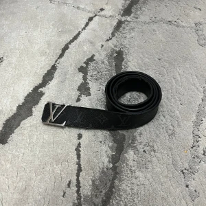 Svart bälte från Louis Vuitton - Snyggt svart bälte från Louis Vuitton med en stilren LV-logotyp i silver på spännet. Bältet har en diskret mönstring och är tillverkat i skinn. Perfekt för att ge en lyxig touch till din outfit. Om ni vill ha några andra bilder eller om ni har frågor är det bara att skriva 