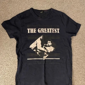 Mörkblå t-shirt med tryck från Jack & Jones - Säljer en mörkblå t-shirt från Jack & Jones med ett coolt tryck av Muhammed Ali och texten 'The Greatest'. Perfekt för dig som gillar sport och stil. T-shirten är kortärmad och gjord i bomull för en bekväm passform. Storlek L men sitter som M skulle jag säga. Hör av er vid funderingar:)