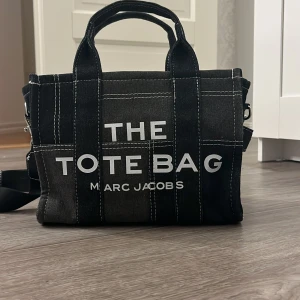 Svart axelväska från Marc Jacobs - Snygg svart axelväska från Marc Jacobs med texten 'THE TOTE BAG' på framsidan. Väskan har ett avtagbart axelband och är perfekt för att bära med sig det nödvändigaste. Den har en stilren design med vita sömmar som ger en cool kontrast.