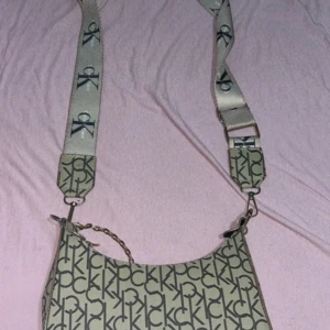 Beige axelväska med CK-logga - Snygg beige axelväska från Calvin Klein med deras ikoniska CK-logga över hela väskan. Väskan har en justerbar axelrem med samma logga och en kedjedetalj som ger en extra touch. Perfekt för att lyfta din outfit!