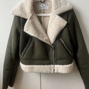 Olivgrön jacka med teddyfoder från Stradivarius - Säljer en snygg olivgrön jacka från Stradivarius med mysigt vitt teddyfoder. Jackan har en cool bikerstil med dragkedjor och bred krage. Perfekt för kyliga dagar när du vill vara både varm och trendig. Använd endast ett fåtal gånger, nästan som ny! Skriv om ni vill ha fler bilder eller har frågor