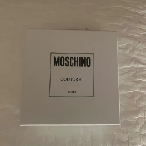 Svart bälte från Moschino - Snyggt svart bälte från Moschino med stora, glittriga guldbokstäver. Perfekt för att ge din outfit en lyxig touch. Kommer i originalförpackning med en skyddande påse. Helt oanvänd. 
