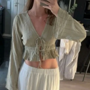 Grön blus med knyt - Säljer en snygg grön blus med knyt framtill och volangdetaljer. Blusen har långa ärmar och en v-ringad design som ger en trendig look. Perfekt  för sommarn och matchar med en vit lång kjol!!
