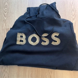 Svart hoodie från BOSS - Säljer en svart hoodie från BOSS med stor logga i guld på framsidan. Tröjan har en känguruficka och justerbar huva med dragsko. Perfekt för en avslappnad stil. Toppskick