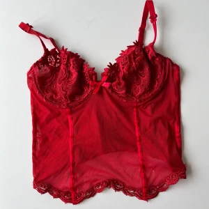 Röd spetskorsett - Snygg röd korsett med spetsdetaljer och justerbara axelband. Den har en vacker blommig spetsdesign och en liten rosett framtill.