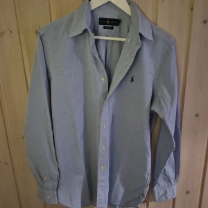 Blå skjorta från Ralph Lauren - Snygg ljusblå skjorta från Ralph Lauren med klassisk passform. Skjortan har långa ärmar och knappar framtill. Den ikoniska loggan är broderad på bröstet. Perfekt för en stilren look.