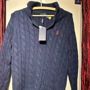 Blå stickad tröja från Polo Ralph Lauren - Sn Säljer en snygg blå stickad tröja från Polo Ralph Lauren med kabelstickat mönster och en dragkedja vid halsen. Tröjan har en röd broderad logga på bröstet och långa ärmar. Perfekt för kyliga dagar! OBS! Priset är inte hugget i sten. Hör av er vid minsta fundering 🍾