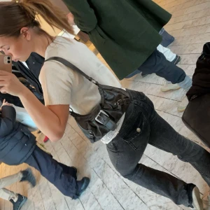 Svart axelväska med spänne - Snygg svart axelväska i skinn med ett stort dekorativt spänne framtill. Väskan har Axel remm och flera detaljer som ger den en cool look. Perfekt för att bära dina nödvändigheter med stil.