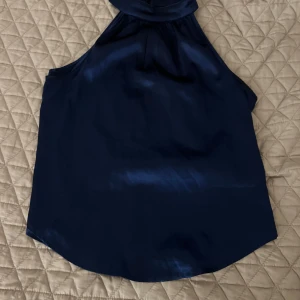 Mörkblå ärmlös topp från SHEIN - Säljer en elegant mörkblå ärmlös topp från  SHEIN. Toppen har en hög hals och är tillverkad i ett glansigt material som ger en lyxig känsla. Perfekt för en stilren look.