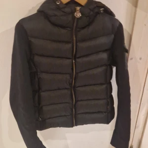 Svart pufferjacka från Moncler - Snygg svart pufferjacka från Moncler med huva och dragkedja framtill. Jackan har långa ärmar och en stilren design med Moncler-logga på ärmen. Perfekt för kyligare dagar och varma. Storlek S.