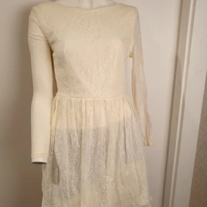 Vit / crémefärgad spetsklänning - Elegant vit spetsklänning med långärmad design och rund hals. Klänningen har en vacker spetsdetalj över hela tyget och en dragkedja i ryggen för enkel påklädning. Perfekt för speciella tillfällen.