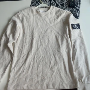 Beige långärmad tröja från Calvin Klein - Säljer en stilren beige långärmad tröja från Calvin Klein. Tröjan har en diskret textur och ett svart märke med CK-loggan på ärmen. Perfekt för en avslappnad och trendig look.