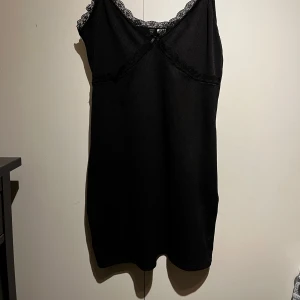 Svart klänning med spetsdetaljer - Elegant svart klänning med spetsdetaljer vid halsringningen och axelbanden. Perfekt för en stilren look. Klänningen har en enkel och klassisk design som passar många tillfällen.