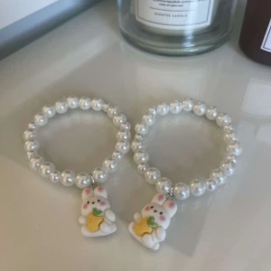 Söta pärlarmband med kaninberlocker - Två supergulliga armband med vita pärlor och små kaninberlocker som håller en morot. Perfekta för att ge en lekfull touch till din outfit. Armbanden är elastiska och passar de flesta handleder. Perfekt accessoar för både vardag och fest!