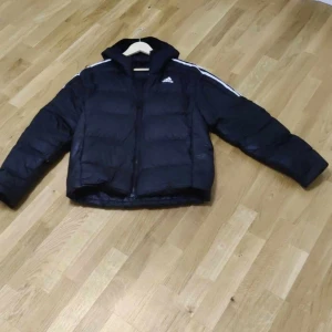 Svart dunjacka från Adidas - Säljer en svart dunjacka från Adidas i storlek M. Jackan har en sportig look med vita detaljer längs ärmarna och en dragkedja framtill. Perfekt för kyliga dagar med sin pufferdesign som håller dig varm. Adidas-loggan syns tydligt på bröstet. Passar både till vardags och sportiga aktiviteter.