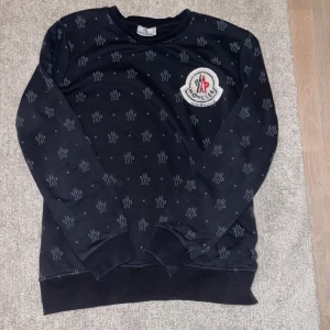 Moncler tröja - Säljer en tvär snygg svart sweatshirt från Moncler med deras ikoniska logga på bröstet. Tröjan har ett diskret mönster över hela ytan och är i mycket bra skick. Perfekt för både vardag och lite kyligare dagar. Passar till allt och är superbekväm! Skriv vid minsta lilla fundering😀✍️✅