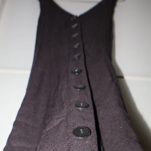 Svart klänning med knappar från Indiska - Snygg svart klänning från Indiska i storlek 40. Den har en djup v-ringning och knappar längs framsidan. Perfekt för både vardag och festliga tillfällen. Klänningen är ärmlös och har en elegant, lång design.