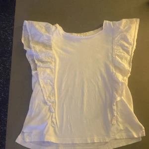 Vit blus med spetsdetaljer - Säljer en söt vit topp med spetsdetaljer på ärmarna. Den har en rund hals och volangdetaljer som ger en romantisk touch. Perfekt för sommardagar eller en avslappnad vårlook. Toppen är i bra skick och passar till både jeans och kjol.
