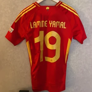 Spanien em 2024 lamine yamal - Säljer en röd fotbollströja från Adidas med gula detaljer och nummer 19. Tröjan har korta ärmar och är i mycket bra skick. Perfekt för fotbollsfans som vill ha en snygg och bekväm tröja att bära på matchdagar!