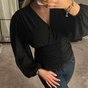 Svart blus med v-ringning - Snygg svart blus med v-ringning och långa puffiga ärmar. Den har en åtsittande midja med rynkade detaljer som ger en smickrande siluett. Perfekt för både vardag och fest!