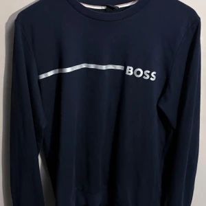 Mörkblå tröja från Boss - Säljer en stilren mörkblå tröja från Boss med ett silvrigt tryck på framsidan. Tröjan har långa ärmar och en klassisk rund halsringning. Perfekt för en avslappnad men ändå snygg look. Passar både till vardags och lite finare tillfällen.