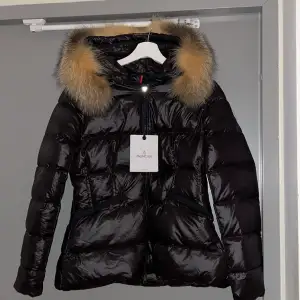 Säljer en snygg svart dunjacka från Moncler i mycket bra skick, aldrig andvänt och har lappar kvar. Jackan har en fluffig äkta pälsdetalj på huvan och en glansig svart färg, en figursydd passform och långa ärmar. Jag kan absolut tänka mig diskutera priset, bara skriva :)