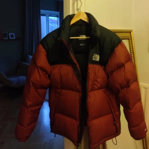 1996 Neptuse puffer från North face - Säljer en snygg röd pufferjacka från The North Face. Den är perfekt för kalla dagar med sin tjocka vaddering och har en dragkedja framtill. Jackan har en klassisk design med svarta detaljer vid axlarna och kragen. Perfekt för vintern! ❄️