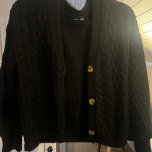 Svart stickad kofta med knappar - Mysig svart stickad kofta med snygga flätmönster och tre knappar framtill. Perfekt för kyliga dagar och att ha över en t-shirt eller blus. Koftan har en klassisk design som passar till det mesta. Ett måste i garderoben för höst och vinter!