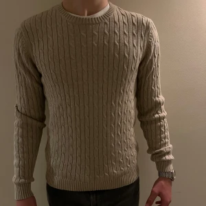 Beige stickad tröja - Säljer en snygg beige stickad tröja med kabelstickat mönster. Perfekt för kyliga dagar och passar både till vardags och lite finare tillfällen. Tröjan har långa ärmar och en rund halsringning. Den är i bra skick och skön.