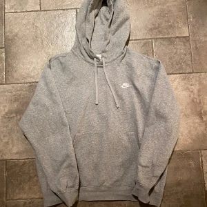 Nike Hoodie Medium - Säljer en klassisk grå hoodie från Nike i storlek M. Sitter stort i storleken. Den är supermjuk och perfekt för både chill och träning. Hoodien har en känguruficka och justerbara snören vid huvan. Nike-loggan är snyggt broderad på bröstet. Perfekt för höst och vår!