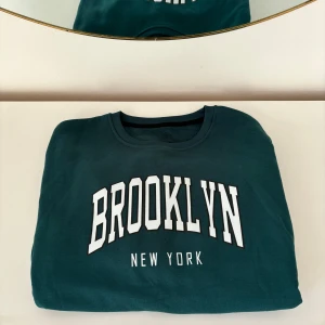 Grön sweatshirt med Brooklyn-tryck - Säljer en snygg grön sweatshirt från STMOVE med texten 'Brooklyn New York' i vitt på framsidan. 