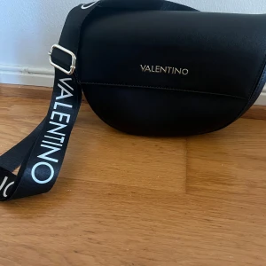 Svart axelväska från Valentino - Snygg svart axelväska från Valentino i skinn. Väskan har en justerbar axelrem med märkeslogga och en stilren design. Perfekt för både vardag och fest. 🖤