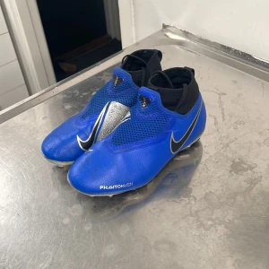 Blå Nike Phantom fotbollsskor - Säljer ett par blå Nike Phantom Vision fotbollsskor i bra skick. De har en snygg design med svarta och silverdetaljer samt en strumpliknande passform för extra komfort. Perfekta för fotbollsträning eller match! ⚽