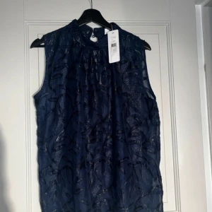 Mörkblå ärmlös topp från Saint Tropez - Snygg mörkblå ärmlös topp från Saint Tropez oanvänd. Den har ett elegant mönster och knytning i nacken. Perfekt för både vardag och festliga tillfällen. Toppen är i storlek L och har en luftig passform. Inköpspris 400kr